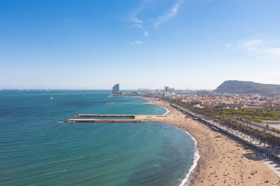 Barcelona's seafront from the air: deep blue sea, a curving beach lined with palms and the city running up to a green hill