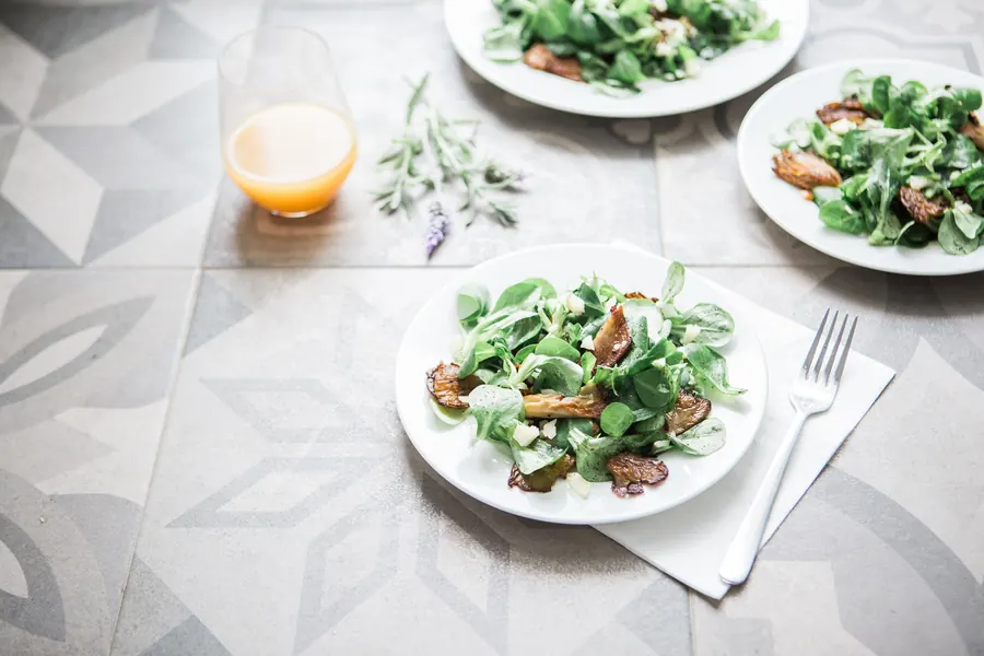 Three plates of leafy salad with roasted vegetables and a glass of juice on patterned floor tiles