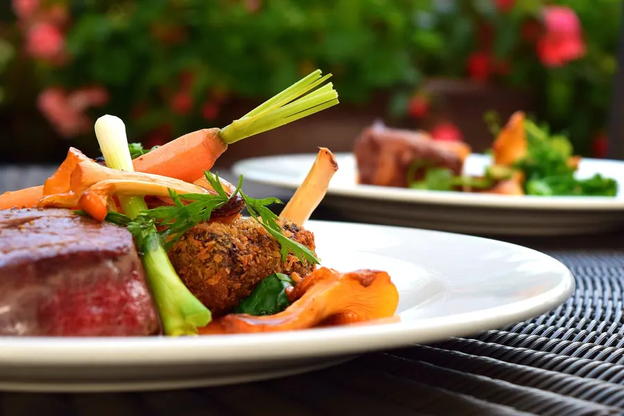 A pink-centred steak with roasted carrots, spring onion and greens on a white plate, a second plate and a green garden behind