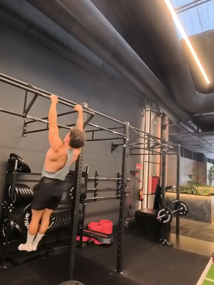 Gio hanging from a wall-mounted pull-up bar next to plate storage in an industrial gym with skylights