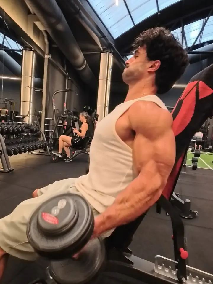 Gio in a cream tank top doing incline dumbbell curls on a bench in a bright gym with a glass roof