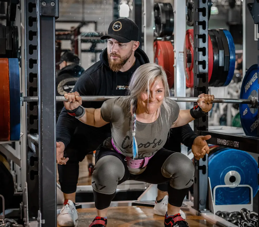 A coach spots a smiling woman as she squats a barbell in a power rack.