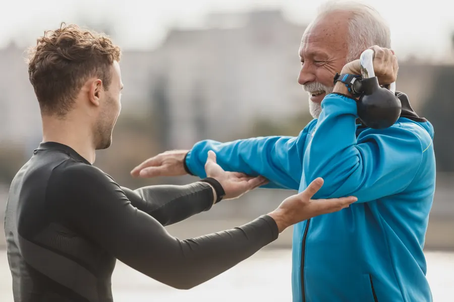 A coach corrects an older man's arm position as he holds a kettlebell outdoors.
