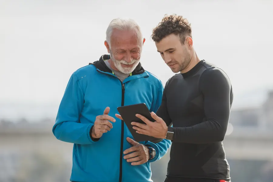 A coach shows an older client his progress on a tablet, both smiling, outdoors.