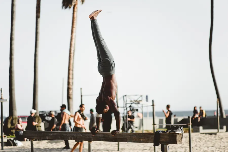 A shirtless athlete holds a handstand on a wooden beam at an outdoor workout area on a sunny beach lined with palm trees
